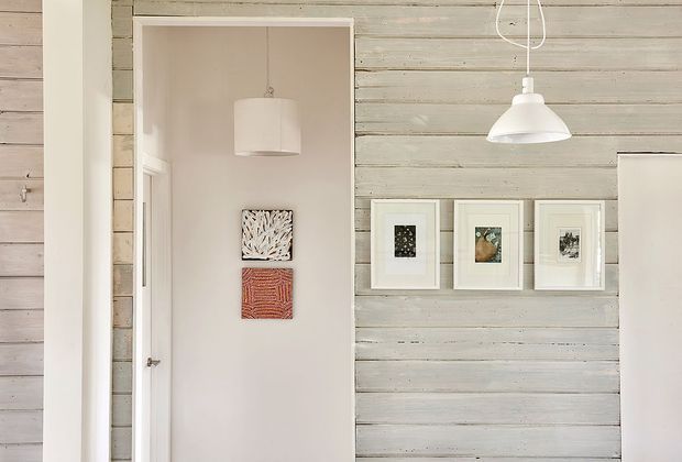 A narrow interior passage or entry area with pale horizontal timber wall panelling, white-painted trim, and a partially open doorway leading into another room. Simple white pendant lights hang from the ceiling, while small framed artworks and textile-style prints decorate the walls. The overall scheme is light, understated, and coastal in character.