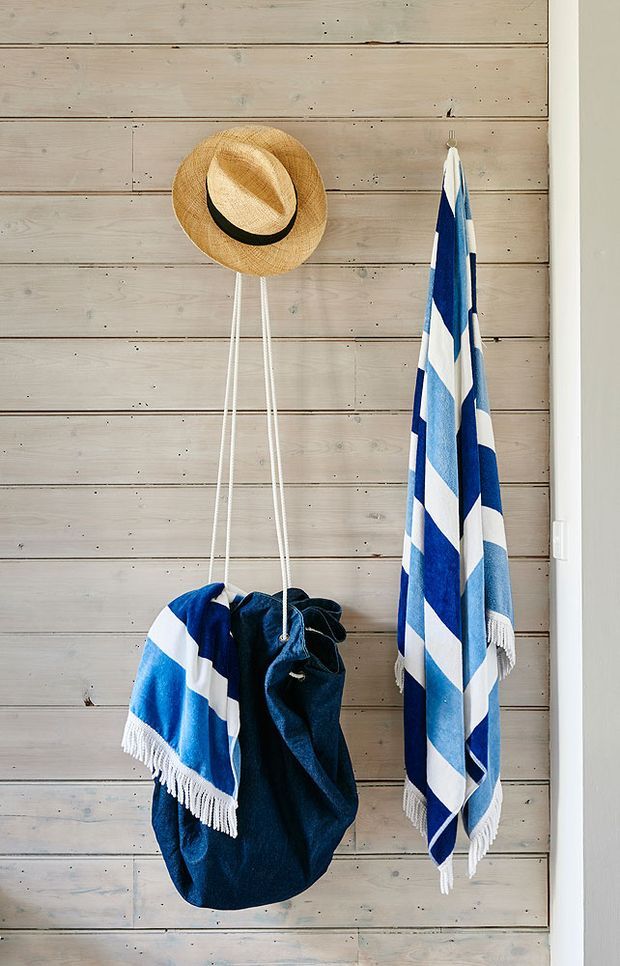 A close-up interior detail of a coastal-inspired wall-mounted storage area. Weathered pale timber boards form the backdrop, with a straw hat, blue-and-white striped beach towel, and dark fabric bag hanging from hooks and cords. The scene has a relaxed beach-house character with strong navy and white accents.