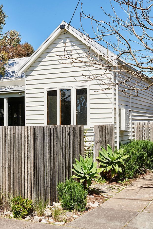 Exterior view of a compact single-storey cottage with white horizontal weatherboard cladding, a prominent pitched gable roof, white-framed windows, and a small covered side entry. A tall vertical timber privacy fence, paved footpath, and low planted garden border the house.