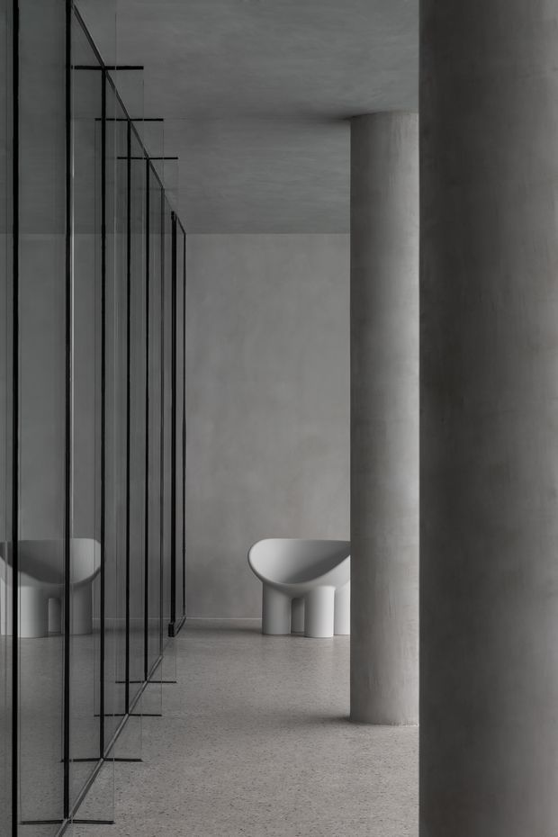 A monochromatic minimalist interior featuring a freestanding sculptural white bathtub, cylindrical concrete columns, pale textured walls and floor, and a long partition of slim black-framed glass panels with reflections. The composition is architectural and gallery-like, with strong vertical lines and a quiet, refined atmosphere.