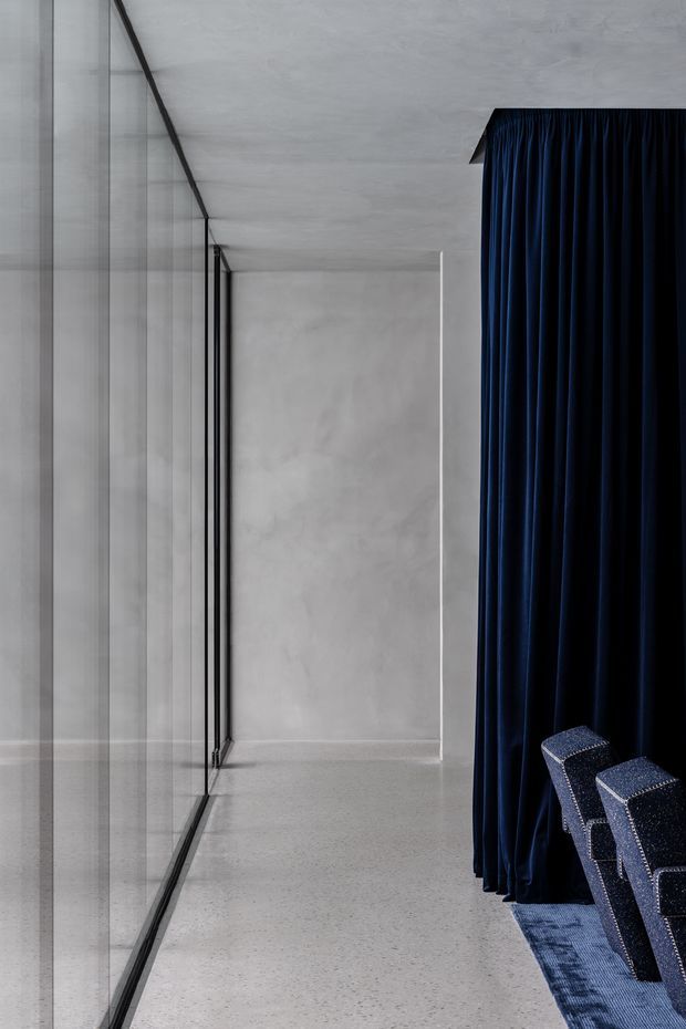 A minimalist interior corridor or circulation space with polished light-grey concrete or terrazzo surfaces, full-height glazing with slender black frames on the left, and a dramatic floor-to-ceiling deep navy curtain on the right. Several dark blue upholstered chairs are partially visible beside a blue patterned carpet, suggesting an adjacent auditorium, screening room, or event space. The composition is highly architectural, with a restrained monochrome palette punctuated by navy blue.
