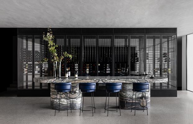 A dramatic contemporary kitchen and home bar featuring a large sculptural stone island with four dark blue bar stools. Behind the island is a full-height black-framed glazed and ribbed screen enclosing dark cabinetry and display shelving. The space has a polished concrete floor, a textured grey ceiling, reflective surfaces, and monochromatic charcoal, grey, and blue tones.