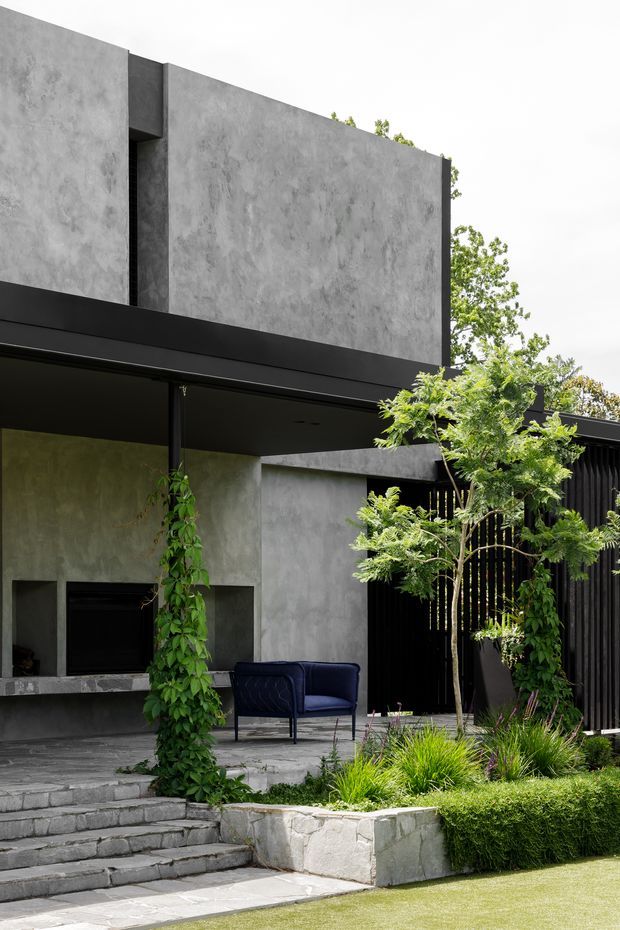 Exterior view of a contemporary multi-level home with a strongly geometric form, dark projecting canopy, textured grey concrete or render walls, black vertical-screen detailing, and a landscaped front terrace. Broad steps lead up to a covered outdoor sitting area with a navy lounge chair, integrated fireplace opening, climbing greenery, ornamental grasses, and a low stone retaining planter.