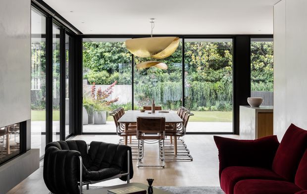 Contemporary open-plan living and dining area with floor-to-ceiling black-framed glazing overlooking a lush garden. A sculptural timber pendant hangs above a timber dining table with upholstered chairs, while a black lounge chair and deep red seating occupy the foreground. A linear fireplace and pale stone or plaster surfaces contribute to the refined, high-contrast interior.