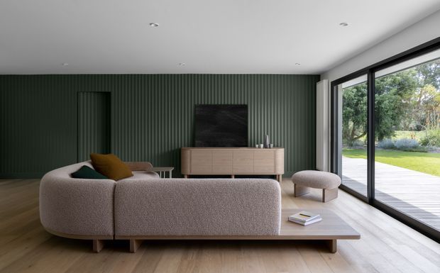 A contemporary living room with a large curved-back neutral sofa, low timber base, and matching ottoman facing a minimalist timber media console. The room features a deep green vertically ribbed feature wall, pale timber flooring, recessed ceiling lights, and expansive black-framed sliding glass doors opening to a deck and landscaped garden. The overall composition is calm, refined, and connected to the outdoors.