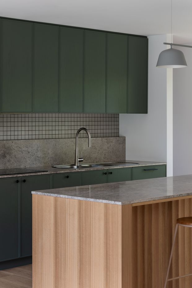 A refined contemporary kitchen with deep green flat-panel cabinetry, a small square-tile splashback, a textured grey stone benchtop and integrated sink, plus a timber island with open seating detail. A sculptural grey pendant light hangs above the work area, creating a muted, sophisticated palette.