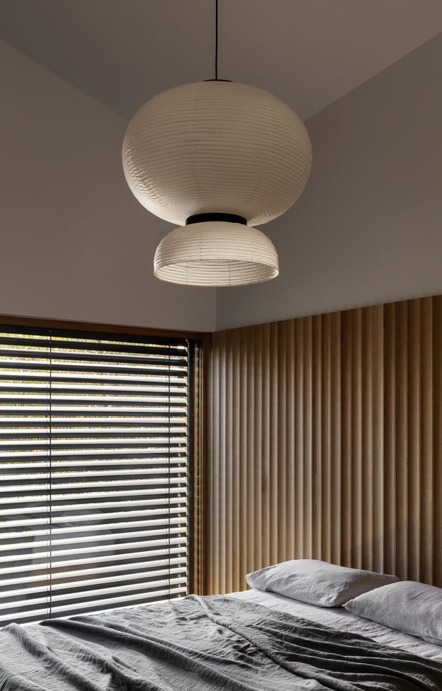 A warm, softly lit bedroom with a low bed dressed in rumpled grey bedding, a full-height vertical timber slat feature wall, and a large window with dark horizontal blinds. Two layered sculptural paper pendant lights hang from the ceiling, creating a calm, tactile interior with strong natural-material detailing.