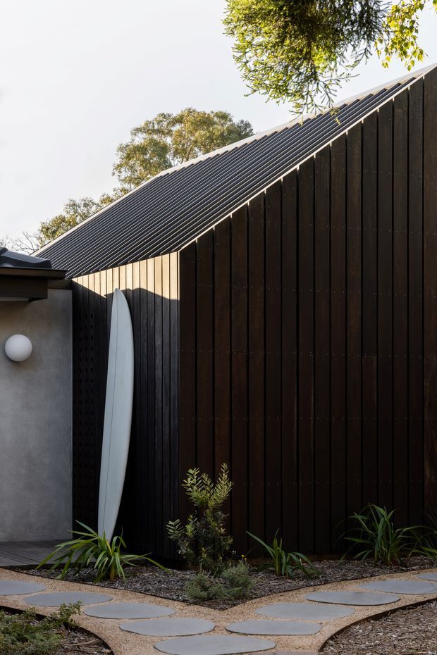 A minimalist landscaped courtyard beside a dark timber-clad building. The tall black-brown vertical timber wall meets a steep corrugated metal gable roof, while a concrete-rendered wall with a round exterior light sits to the left. A white surfboard leans against the wall, and low drought-tolerant planting surrounds irregular circular concrete stepping stones set in gravel.