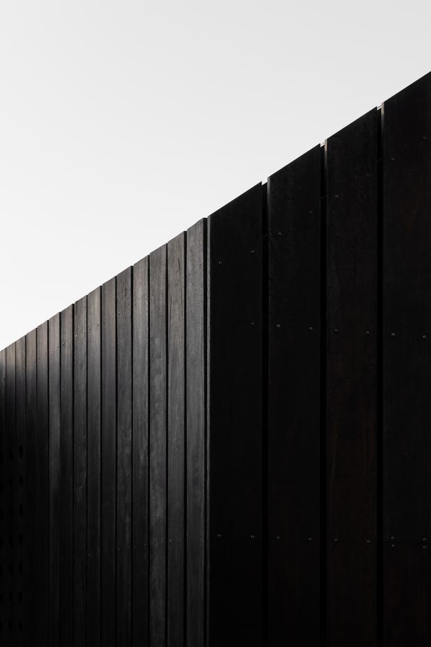 Minimal close-up architectural detail of a tall, dark vertical timber fence or screen. Black-stained boards with visible grain and evenly spaced fasteners form a strong diagonal perspective against a plain pale sky, creating a stark, graphic composition. No interior space or broader building context is visible.
