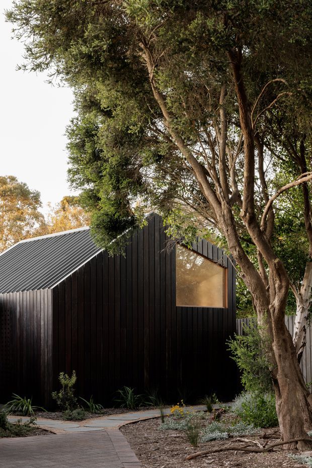 A compact dark-clad building nestled among mature trees and landscaped planting. The exterior has vertical black timber cladding, a steeply pitched corrugated metal roof, and a large warm-toned window. A paved garden path approaches the structure, creating a sheltered, wooded setting.
