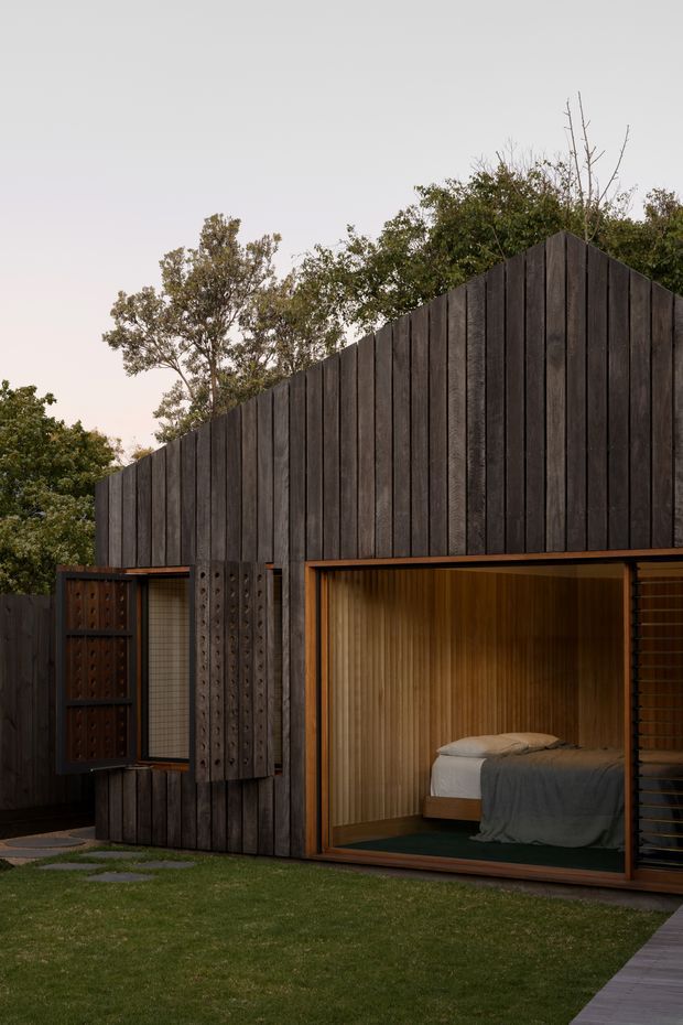 A compact, dark timber-clad garden dwelling with a steep gabled roofline and large glazed opening. The interior visibly contains a bed, while a smaller side opening has timber shutters and patterned screening. Warm timber lining and bronze-toned frames contrast with the charcoal vertical cladding. The structure sits in a landscaped lawn and is surrounded by mature trees in soft evening light.
