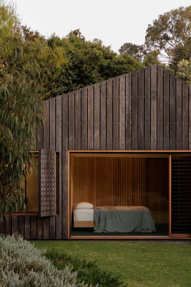 Exterior view of a compact, dark-clad garden room or dwelling set within landscaped greenery. Vertical dark timber cladding forms a steep gable-like facade, with a large glazed opening revealing a simply furnished bedroom inside. Warm timber reveals and interior panelling contrast with the dark exterior, while a lawn, shrubs, and mature trees surround the structure.
