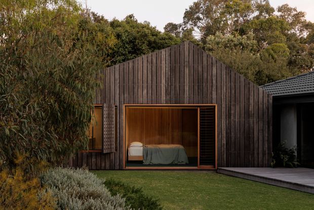 A dark timber-clad gabled pavilion set within a landscaped garden. The facade features a large timber-framed opening revealing a compact bedroom with a bed, warm timber-lined interior, and a dark slatted screen. A paved deck connects to a partially visible dark-roofed structure on the right, with mature planting surrounding the building.