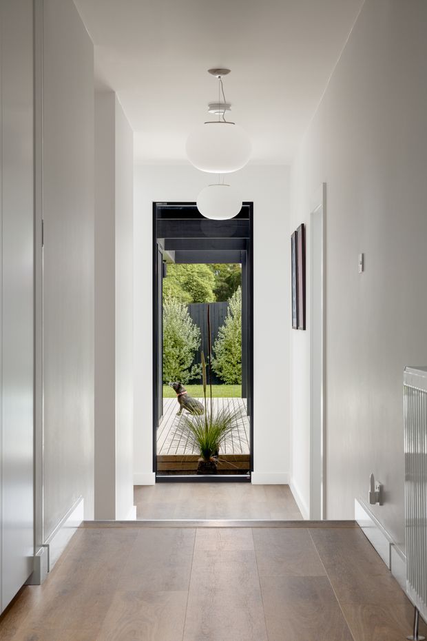 Bright, minimalist residential hallway with white walls, pale timber flooring, recessed doorways and contemporary spherical pendant lights. A black-framed glazed door at the end opens onto a small landscaped outdoor area, while a radiator and framed artwork add functional and decorative details.