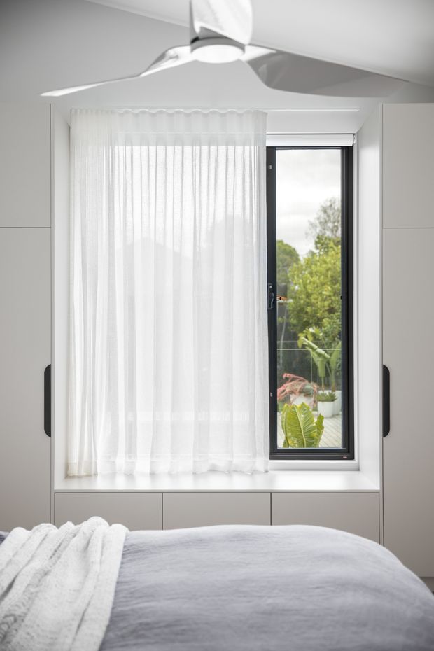 Bright, minimalist bedroom detail focused on a built-in window seat and cabinetry beneath a tall black-framed window. Sheer white curtains soften the daylight, while a white ceiling fan, pale walls, and neatly made bed create a clean, serene interior. Greenery is visible outside through the window.