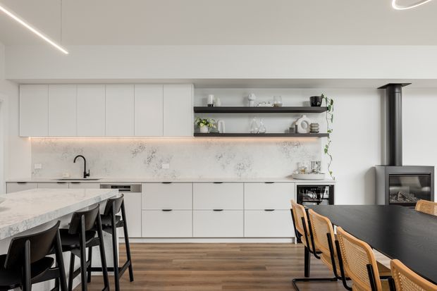 Contemporary open-plan kitchen and dining area with white handleless cabinetry, a pale veined stone splashback and benchtop, open timber shelving, integrated under-cabinet lighting, dark dining furniture, and a black wood-burning fireplace. Warm timber flooring contrasts with the light cabinetry and minimalist linear lighting.