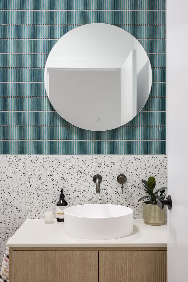A contemporary bathroom vanity featuring a round white vessel basin, wall-mounted brushed metal tapware, timber-look cabinetry, and a large circular mirror. The wall combines white terrazzo-style panels with vertically patterned teal tiles, creating a clean, graphic and refined finish. A small potted plant and bathroom accessories sit on the countertop.