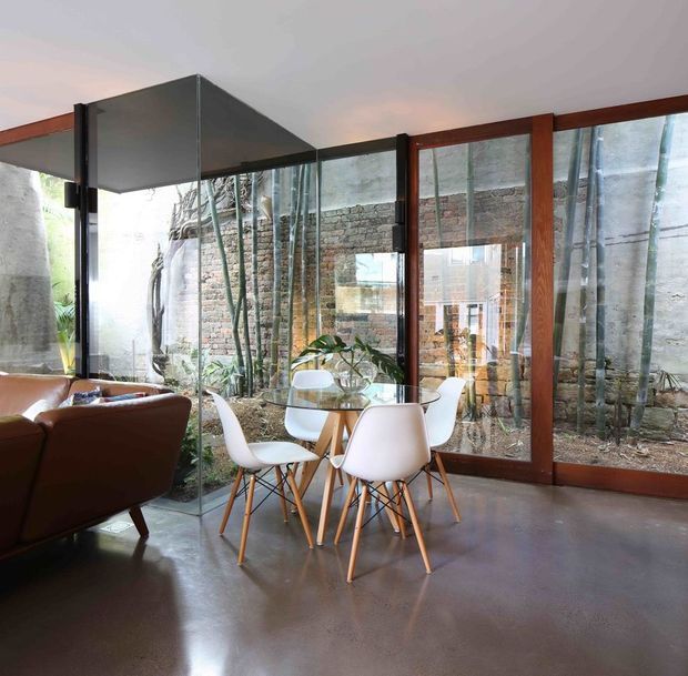 Contemporary open-plan dining and living area with a round glass dining table and white molded chairs beside a brown leather sofa. Full-height glazing and timber-framed doors overlook a planted courtyard and exposed brick boundary wall, while a dark polished concrete floor and angular suspended ceiling feature create an industrial-modern character.