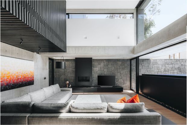 Double-height contemporary living room with a large light-grey sectional sofa facing a black built-in media wall and television. Rough grey stone forms the feature wall, with exposed concrete beams and a dramatic black ribbed ceiling element. Floor-to-ceiling glazing brings in natural light, and colourful cushions and abstract artwork provide small accents.