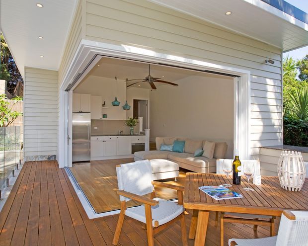 Bright coastal-style outdoor living and dining deck opening directly into a compact open-plan kitchen and living room. The space features white horizontal weatherboard cladding, wide sliding/folding glazed doors, timber decking and flooring, white cabinetry, a stainless-steel refrigerator, a beige sofa with turquoise cushions, and a timber dining table with white outdoor chairs. Recessed ceiling lights and a ceiling fan support a relaxed indoor-outdoor connection.