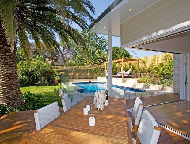 A bright contemporary outdoor dining area on a timber deck, opening directly to a landscaped backyard with a swimming pool. The space features a large timber dining table, white woven dining chairs, frameless glass balustrades, and a white-clad house with broad open glazing and recessed ceiling lights. Palm trees, garden planting, and a timber pergola create a resort-like setting.
