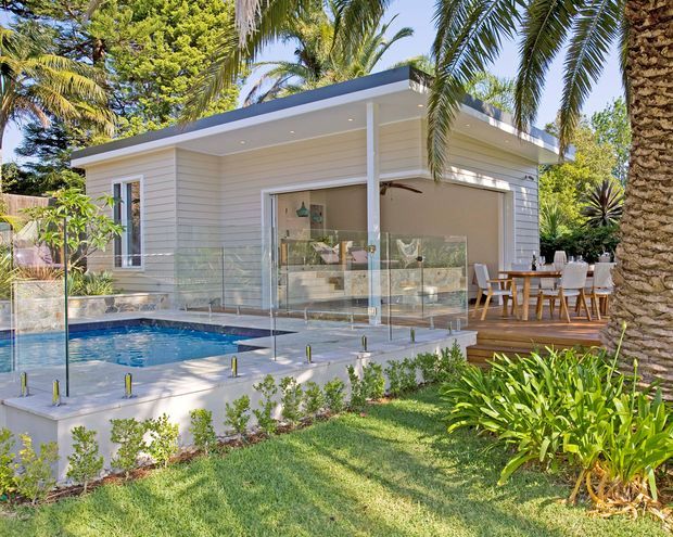 A bright single-storey pavilion-style home surrounded by tropical landscaping, with pale horizontal weatherboard cladding, a low flat roof, and large open glazing. The image focuses on an outdoor pool terrace enclosed by frameless glass balustrades, with a timber deck and dining area beside the building. Palm trees and lush planting frame the pool and lawn.