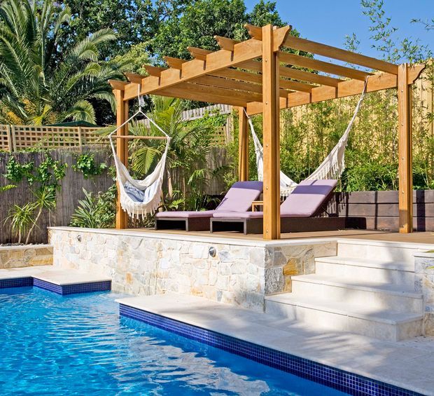 A bright landscaped poolside outdoor living area with a timber pergola, hanging hammocks, and two cushioned loungers on a raised stone terrace. Pale stone retaining walls and steps lead down to a swimming pool finished with a dark blue tiled waterline. Timber screening, dense tropical planting, and palms enclose the private space.
