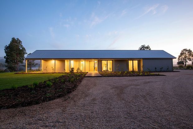Single-storey contemporary rural residence photographed at dusk, with a long low rectangular form, symmetrical pitched metal roof, dark grey exterior walls, extensive warm-lit glazing, a covered front verandah, landscaped garden beds, and a broad gravel driveway. Mature trees and open grounds surround the house.