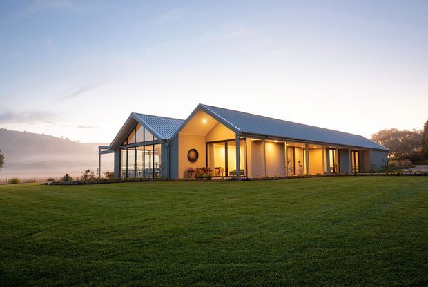 A single-storey contemporary rural house photographed at dusk, set within a broad manicured lawn. The building has a long low gabled form with dark metal pitched roofs, pale vertical wall cladding, extensive floor-to-ceiling glazing, and a prominent glazed gable end. Warm interior lighting spills onto a covered outdoor terrace, creating a welcoming contrast with the cool evening landscape.