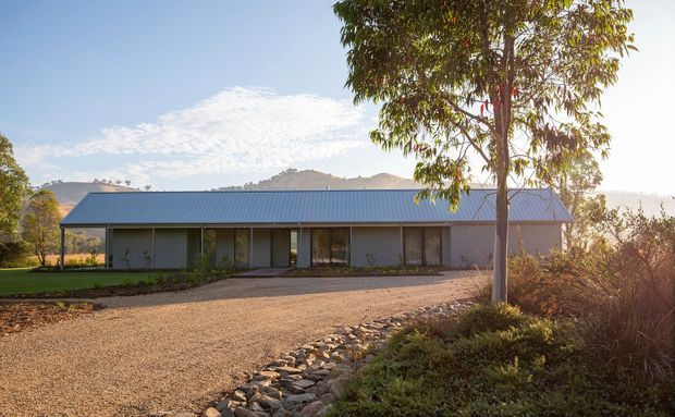 Single-storey elongated residential building set in a landscaped rural or semi-rural setting. The house has a simple low-pitched gable roof with grey metal roofing, a restrained horizontal facade, large dark-framed glazed openings, and muted grey wall cladding. A broad gravel driveway and dry-stone drainage edge lead toward the house, with mature planting and distant hills behind it. The image has warm late-afternoon light and a contemporary, understated character.