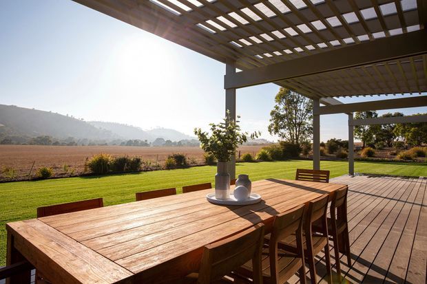 A spacious outdoor dining terrace with a large solid timber dining table and matching chairs beneath a timber pergola. The deck overlooks a manicured lawn and expansive rural fields, with low hills and trees in the distance. Strong sunlight filters through the open pergola roof, creating a warm, relaxed indoor-outdoor setting.