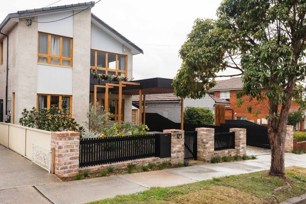 Street-facing view of a contemporary two-storey residential home with a light rendered facade, dark tiled pitched roof, warm timber-framed windows, timber pergola, and a black metal front fence set between exposed brick pillars. A neighbouring brick house and mature street tree are visible.