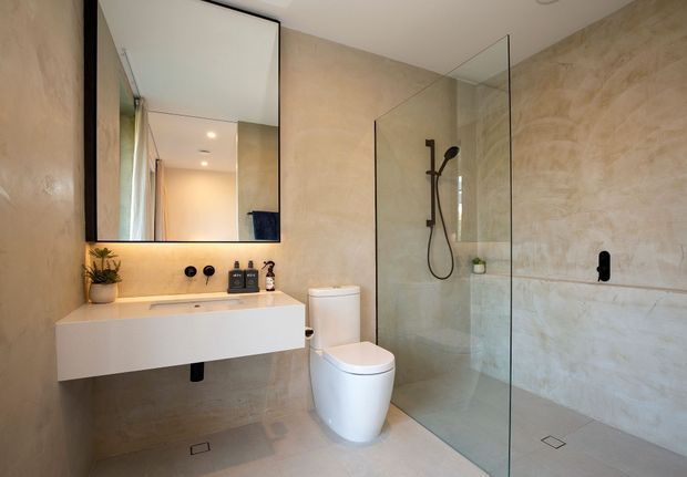 A contemporary bathroom with a wall-mounted white vanity, integrated basin, black tapware and fixtures, frameless glass shower enclosure, wall-hung mirror, and white toilet. Large beige stone-look tiles cover the walls and floor, creating a warm, minimalist space.