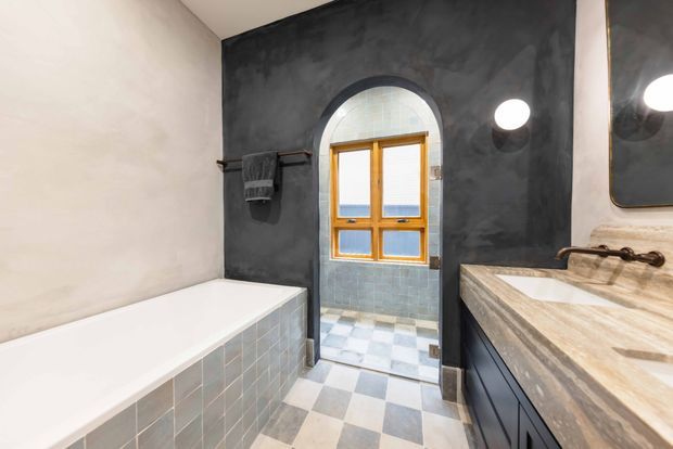 A moody, characterful bathroom with a white built-in bathtub clad in pale blue-grey square tiles, a dark textured plaster feature wall, and a wood-framed arched window opening into a tiled area. The room includes a dark vanity with a substantial natural-stone countertop, wall-mounted round lighting, a frameless mirror, and a checkerboard floor in pale grey and off-white tones.