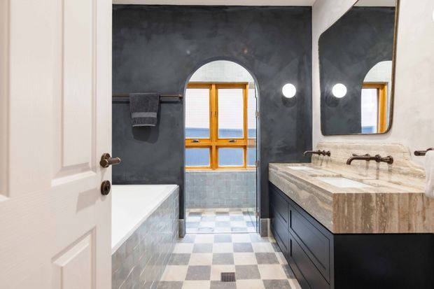 A stylish bathroom viewed through an open white paneled door. The room features a dark charcoal textured wall finish, an arched opening with timber-framed glazing, a freestanding or built-in white bathtub, pale wall tiles, a black and light grey checkerboard floor, and a dark built-in vanity with a thick beige-grey stone benchtop and twin aged-metal wall-mounted taps. A large rounded-corner mirror and circular wall lights add visual interest.