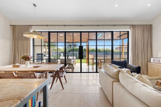 Bright open-plan living and dining area with a large stone kitchen island in the foreground, timber dining table and chairs, a neutral sectional sofa, and full-height black-framed glazing opening to a covered outdoor area. Light curtains, pale walls, and a woven pendant create a warm contemporary interior.