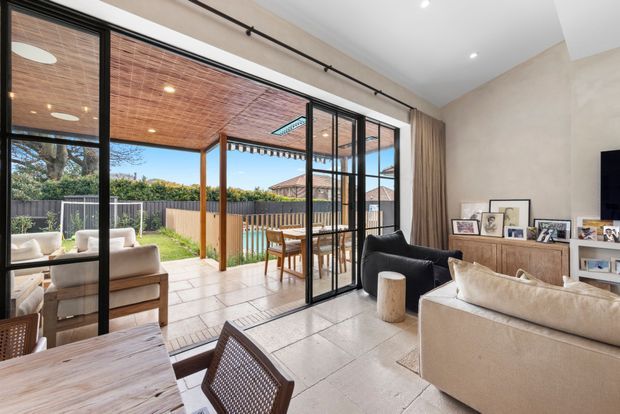 Bright contemporary living room opening through large black-framed glazed sliding doors to a covered outdoor patio and garden. The room features pale tiled flooring, a neutral upholstered sofa and armchairs, a dark lounge chair, timber furniture, built-in timber-toned cabinetry, recessed ceiling lights, and full-height curtains. The outdoor area has a timber-lined ceiling, posts, dining table, and privacy fencing.