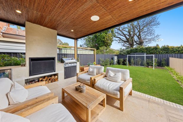 A covered outdoor entertaining area with a timber-lined ceiling, built-in fireplace, outdoor kitchen and grill, timber-framed lounge seating, and a coffee table. The space opens onto a neatly landscaped lawn enclosed by dark fencing.
