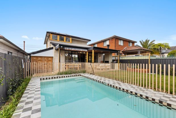 Rear exterior view of a contemporary residential home with a rectangular swimming pool in the foreground, surrounded by a checkerboard-tiled coping and timber slat fencing. The house has dark-framed glazing, a covered outdoor entertaining area, timber and rendered walls, and a landscaped suburban backyard. Neighbouring suburban homes are visible beyond the property.