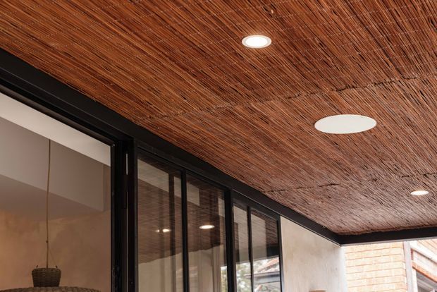 Close-up architectural view of a covered outdoor or semi-enclosed area featuring a richly textured woven timber or reed ceiling, recessed and flush-mounted circular lights, black-framed floor-to-ceiling glazing, and plaster and brick wall surfaces. The space has a warm, natural, contemporary character.
