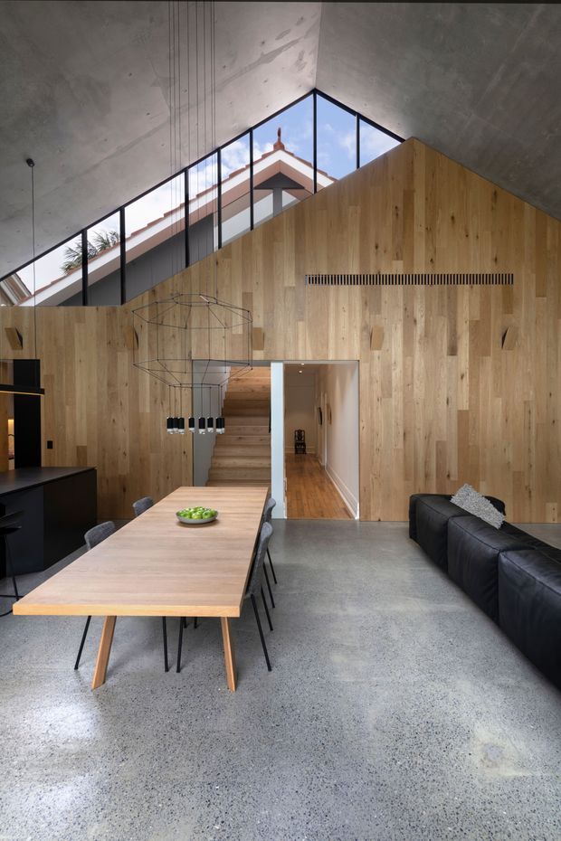 A contemporary open-plan dining and living area with a long timber dining table, black chairs, black kitchen joinery, and a large dark sectional sofa. The room features a dramatic pitched concrete ceiling, full-height black-framed clerestory glazing, extensive vertical timber wall panelling, a timber stair, and a polished speckled concrete floor. A roofline and palm foliage are visible through the glazing.