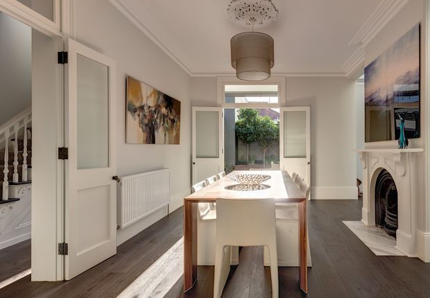 A bright, refined dining room in a traditional-style home, centred on a rectangular timber dining table with white upholstered chairs. The room features dark timber flooring, pale walls, ornate ceiling cornices, a decorative ceiling light, a white-painted fireplace surround, glazed internal doors, and French doors opening to a small courtyard or garden. A staircase and artwork are visible at the left.