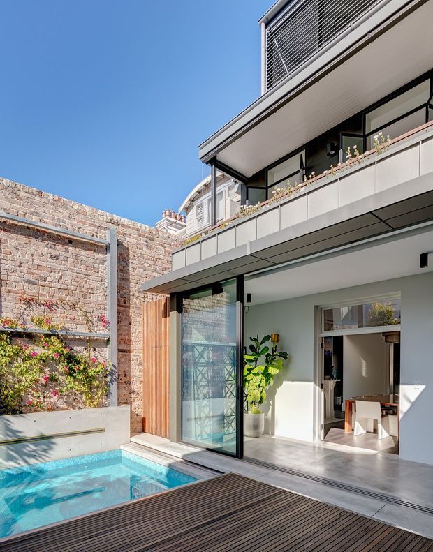 A bright contemporary multi-level residential extension viewed from a private courtyard. The image shows a turquoise plunge pool beside a dark timber deck, exposed brick boundary walls with climbing plants, and a modern white-and-grey facade with extensive glazing, a projecting balcony, louvred upper-level screening, and a large open sliding door into the interior.