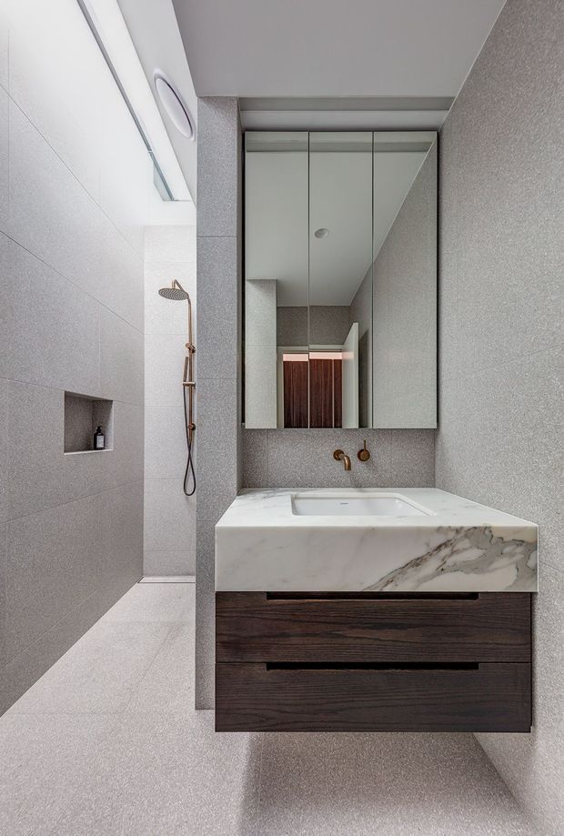 A compact contemporary bathroom with a walk-in shower, wall-mounted dark timber vanity, integrated white marble basin benchtop, brushed brass wall-mounted tapware, and mirrored upper cabinetry. The room is finished in continuous light grey textured wall and floor tiles, with a recessed shower niche and concealed linear ceiling lighting.