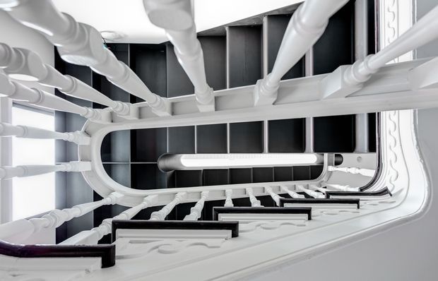 Upward-looking view through a tall, ornate stairwell or atrium, with repeating white decorative balusters and moulded railings surrounding dark recessed wall panels. The dramatic vertical perspective emphasizes the height and symmetry of the interior.
