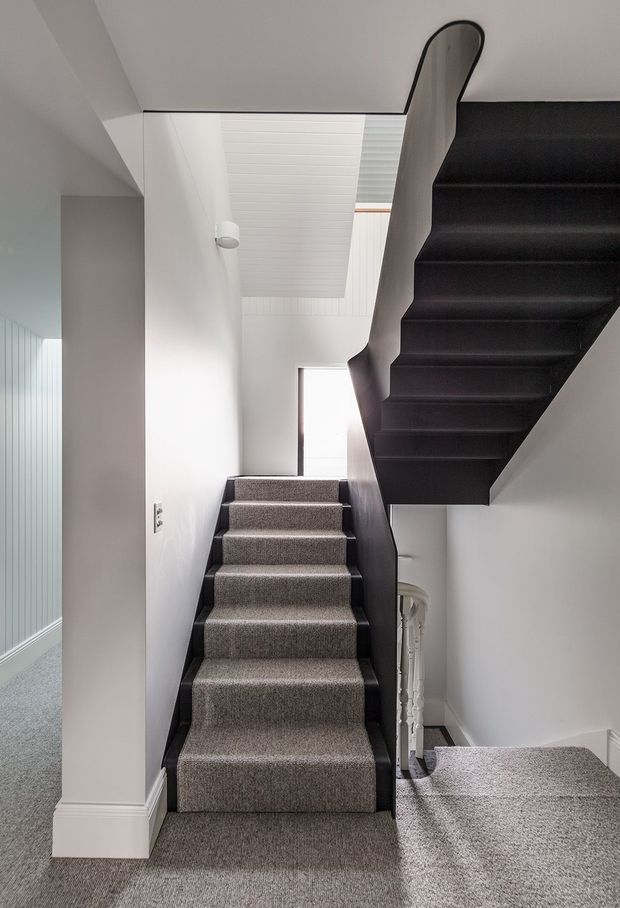 A narrow, bright interior stairway with grey carpeted lower stairs and a contrasting black open-riser staircase ascending overhead. White walls and ceiling, a skylight or roof window, simple white trim, and a small glimpse of a decorative balustrade create a minimalist, high-contrast composition.