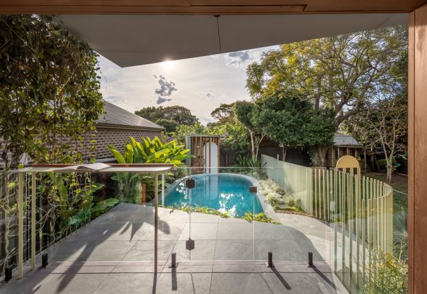 View from a covered outdoor terrace toward a compact turquoise swimming pool, with pale tiled paving, transparent glass balustrades, lush established planting, and surrounding low-rise residential structures. Timber framing and soffit details create a warm contemporary indoor-outdoor connection.