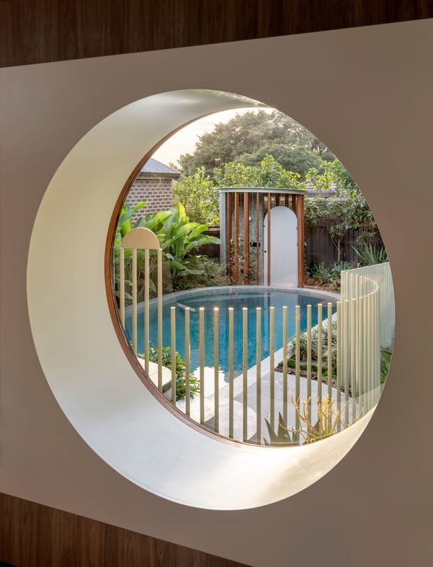 A sculptural circular opening in a pale plaster wall frames a landscaped backyard courtyard with a blue swimming pool, curved pale paving, lush planting, timber screening, and a small arched garden gate. Vertical metal balustrade bars appear in the foreground, while dark timber wall cladding surrounds the opening. The image emphasizes a carefully composed architectural view from inside toward the outdoor pool area.