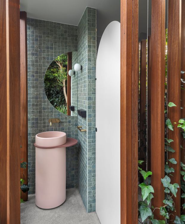 A compact, highly designed powder room viewed through a timber slatted entrance. The room features green square mosaic-tiled walls, a curved tiled partition with an arched opening to lush planting, a large round mirror, and a sculptural pink cylindrical pedestal basin with matching curved benchtop. Brass tapware and fittings add a warm accent, while grey flooring and a curved white wall create contrast. Vertical timber framing and climbing greenery are visible at the entry.