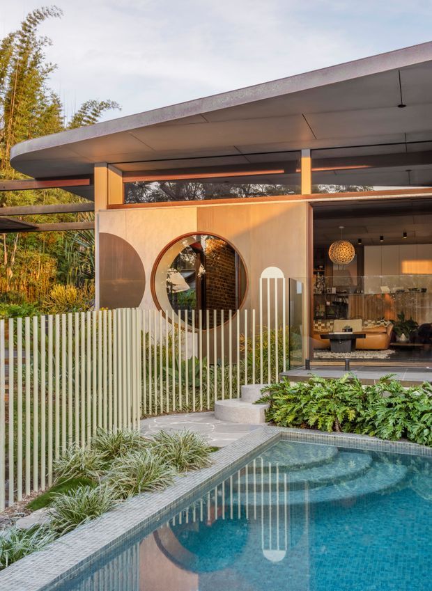 A contemporary single-storey house viewed from the landscaped pool area, featuring a broad low-slung flat roof with deep overhangs, extensive glazing, warm timber-toned wall panels, and distinctive circular window detailing. A slender vertical white picket-style fence borders the tiled swimming pool, with lush planting and curved concrete steps leading to the open living area. The interior visible through the glazing includes warm timber finishes, patterned tile details, pendant lighting, and seating.